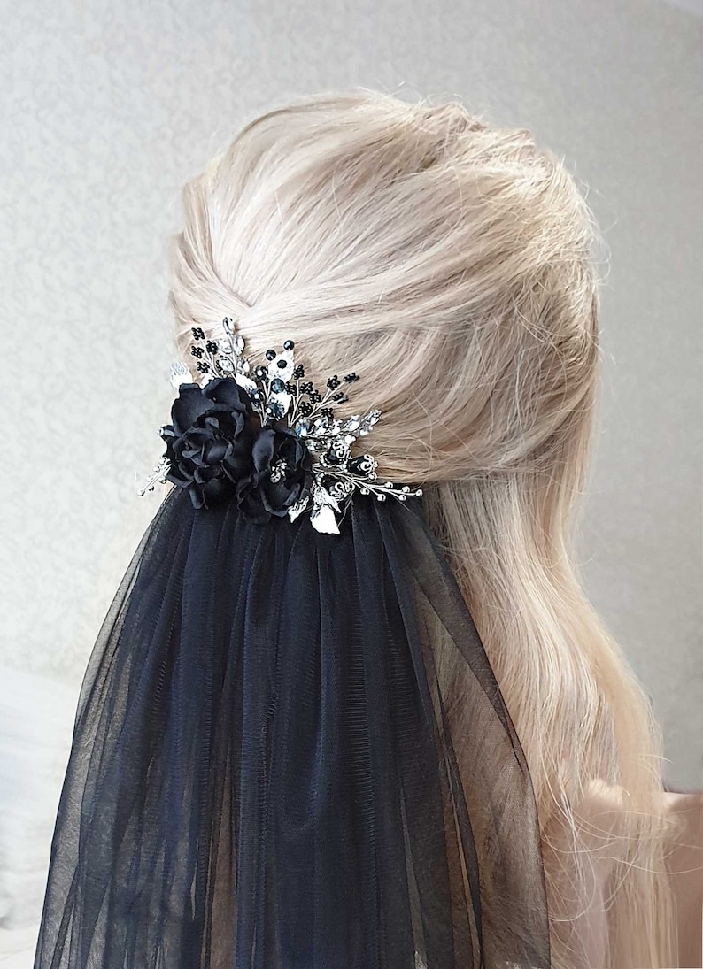 Puede incluir: Un peine para el cabello floral negro con una velo a juego. El peine presenta dos flores de tela, hojas plateadas y detalles brillantes. El velo est&aacute; hecho de una tela negra transparente. El accesorio para el cabello est&aacute; sobre cabello rubio.