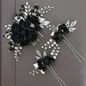 Puede incluir: Un conjunto de tres accesorios para el cabello negros. El conjunto incluye un peine con flores de tela negra y detalles plateados, y dos horquillas con flores negras, hojas plateadas y detalles de cristal y perlas. Los accesorios est&aacute;n sobre una superficie gris oscuro.