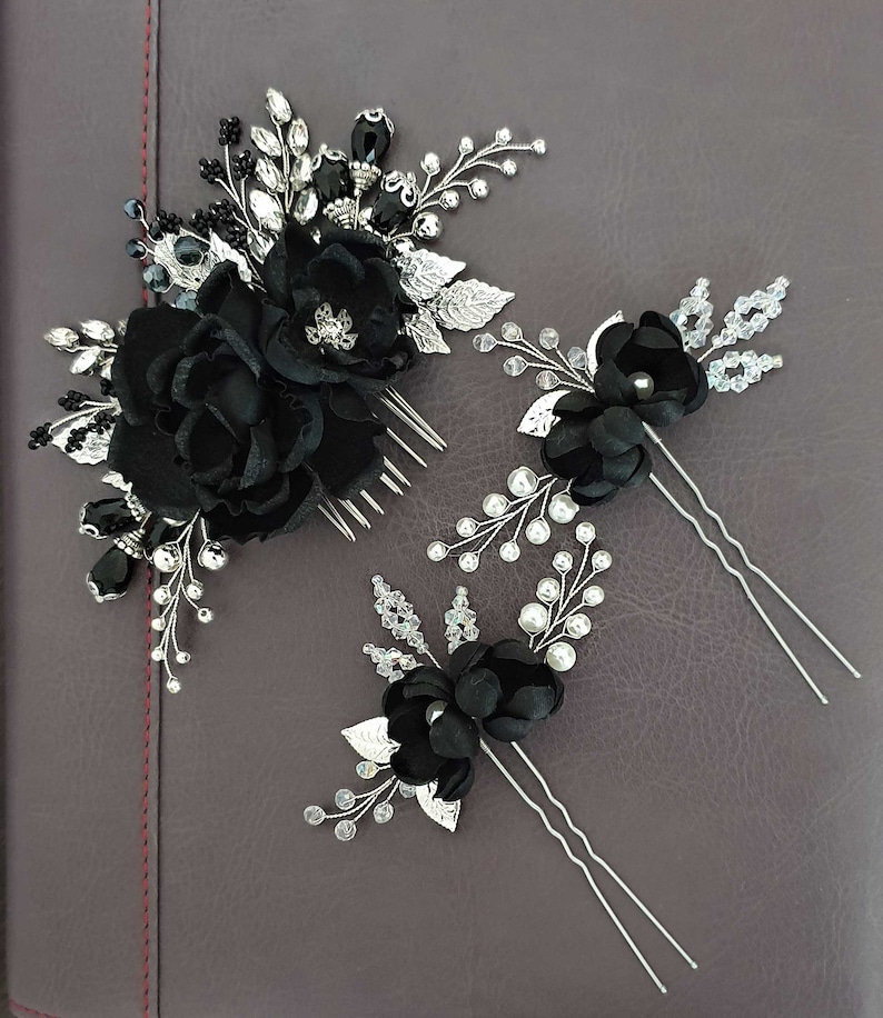 Puede incluir: Un conjunto de tres accesorios para el cabello negros. El conjunto incluye un peine con flores de tela negra y detalles plateados, y dos horquillas con flores negras, hojas plateadas y detalles de cristal y perlas. Los accesorios est&aacute;n sobre una superficie gris oscuro.