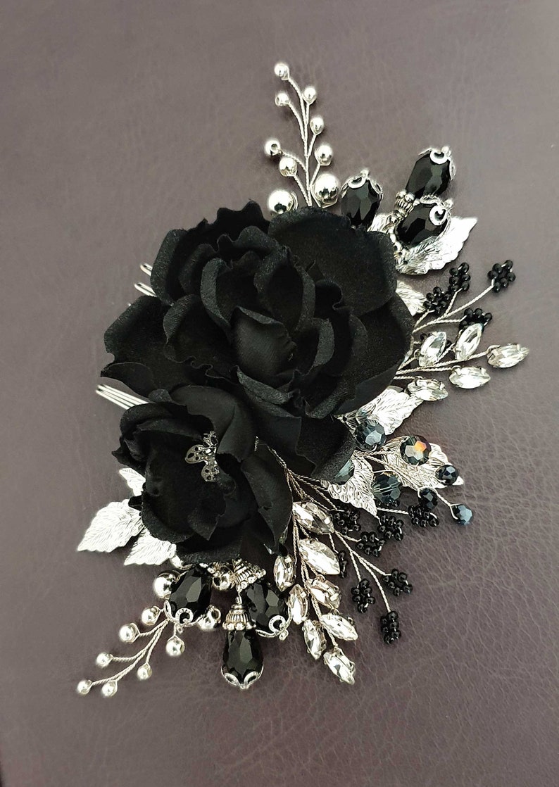 Puede incluir: Una horquilla floral negra con una gran flor de tela y flores de acento m&aacute;s peque&ntilde;as. La horquilla presenta hojas plateadas, cristales transparentes y negros y peque&ntilde;as cuentas negras. Un accesorio elegante para ocasiones especiales.