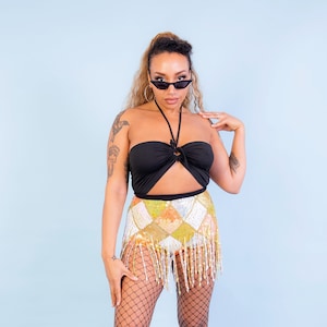 May include: A black halter top with a keyhole detail and a sequined mini skirt with a fringe hem. The skirt features a patchwork design in yellow, orange, and silver. The outfit is completed with black fishnet stockings and black boots.