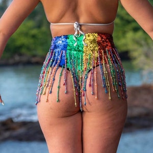 May include: A woman wearing a rainbow sequined bikini bottom with fringe. She is standing on a wooden dock overlooking the ocean.