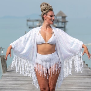 May include: A woman wearing a white bikini and a white fringed kimono stands on a wooden pier. The background features a calm sea and a small structure on the water. The outfit is perfect for a beach or resort setting.