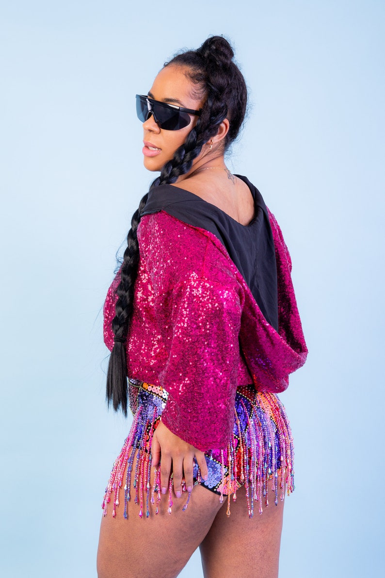 May include: A magenta sequined hooded jacket with a black hood and matching sequined shorts. The shorts feature a colorful beaded fringe. The person is wearing black sunglasses and has long black braids.