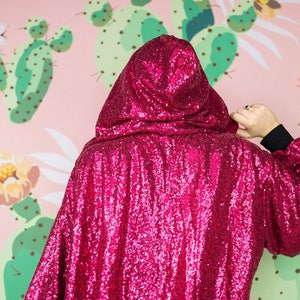 May include: A hooded cape covered in bright pink sequins. The cape is worn over a black long-sleeved shirt. The background features a pink wall with a cactus and floral pattern.