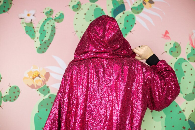 May include: A hooded cape covered in bright pink sequins. The cape is worn over a black long-sleeved shirt. The background features a pink wall with a cactus and floral pattern.