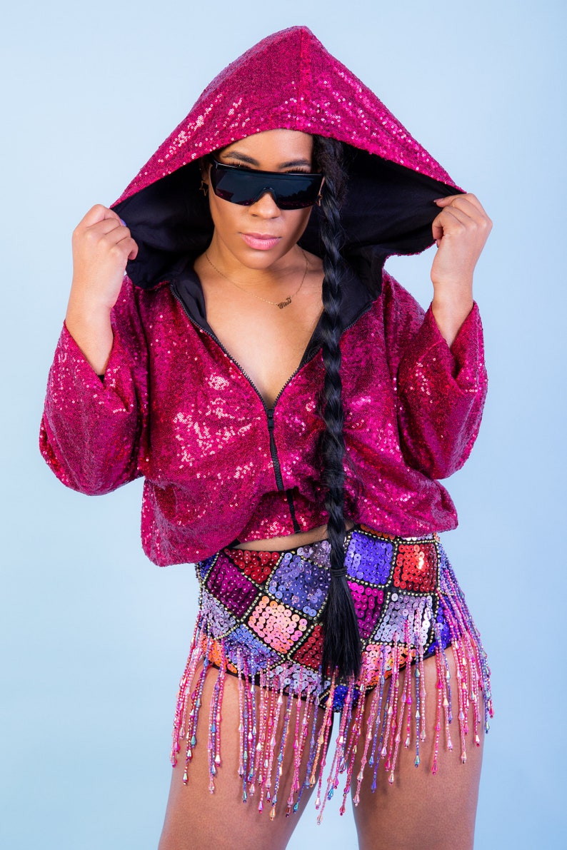 May include: A magenta sequined hooded jacket with a black interior and matching high-waisted shorts. The shorts feature a patchwork design with colorful sequins and beaded fringe. The model is wearing black sunglasses.