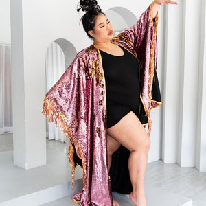 May include: A sequined kimono in shades of pink and gold with fringe trim. The open-front robe is worn over a black outfit. The garment's sleeves are wide and flowing, and the overall style is glamorous and eye-catching.