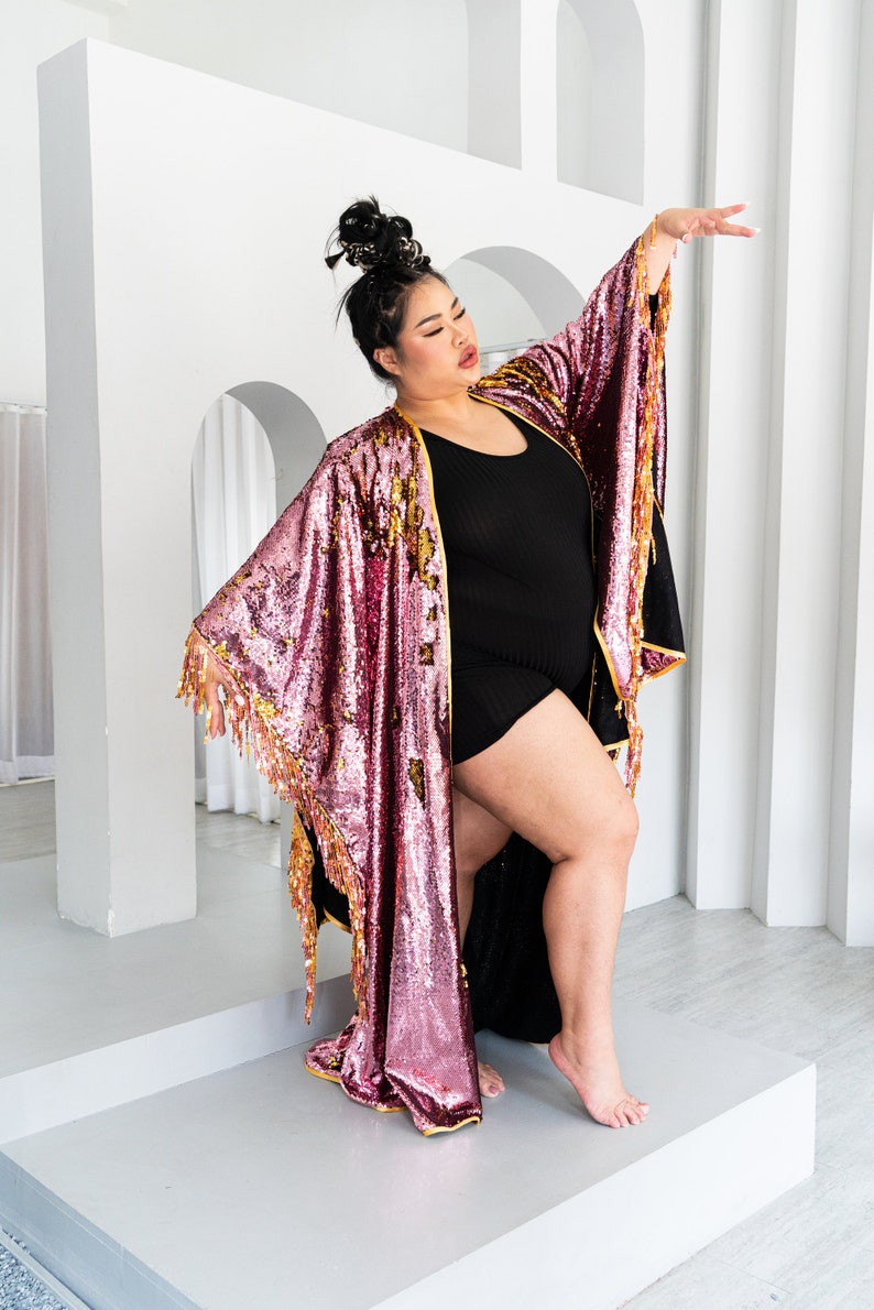 May include: A sequined kimono in shades of pink and gold with fringe trim. The open-front robe is worn over a black outfit. The garment's sleeves are wide and flowing, and the overall style is glamorous and eye-catching.