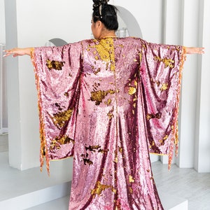 May include: A pink sequined kimono-style robe with gold accents and fringe trim. The robe has wide sleeves and a floor-length design. The gold accents are scattered across the pink fabric. The model's back is facing the camera.