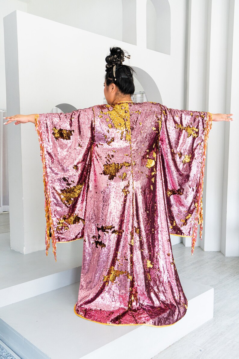 May include: A pink sequined kimono-style robe with gold accents and fringe trim. The robe has wide sleeves and a floor-length design. The gold accents are scattered across the pink fabric. The model's back is facing the camera.