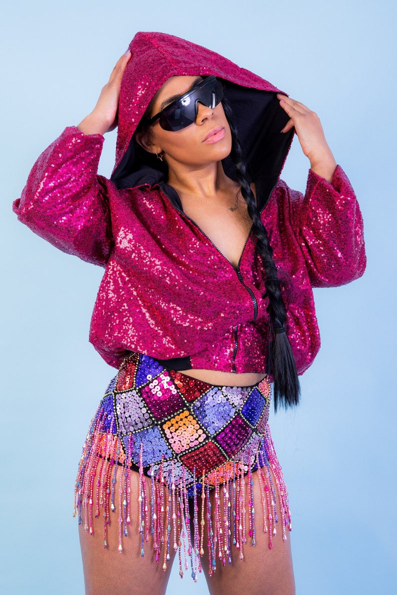 May include: A magenta sequined hooded jacket with a black interior is paired with a matching sequined patchwork mini skirt. The skirt features beaded fringe. The model is wearing black sunglasses.
