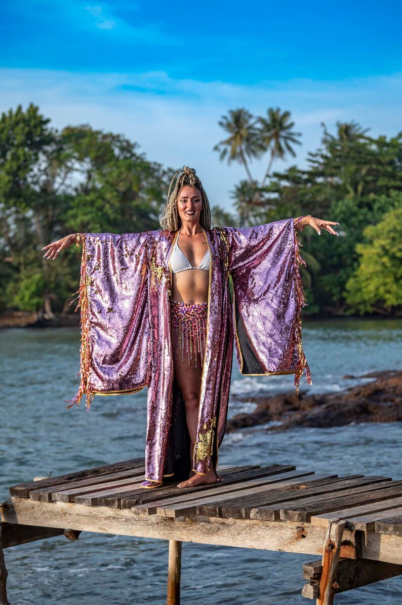 May include: A woman wearing a shimmering, long, open-front robe with large sequins in shades of purple and gold. The robe has long sleeves with fringe and gold trim. She is standing on a wooden dock near the water.