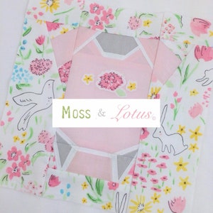 Könnte beinhalten: Ein rosa-grauer Stoffblock mit einem Blumenmuster und einem weißen Vogel- und Hasendesign. Der Block hat den Text "Moss & Lotus" in Grün.