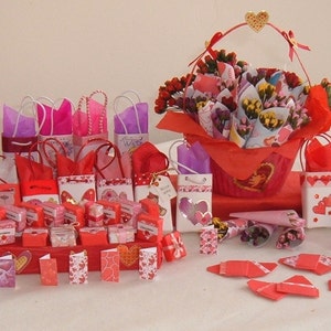 May include: A collection of small red and pink gift bags and boxes decorated with hearts, flowers, and polka dots. The bags and boxes are arranged on a white surface with a basket of flowers in the center.