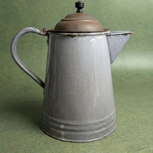 May include: A vintage gray enamel coffee pot featuring a tapered body, a curved handle, and a matching lid with a small black knob. The metal surface shows signs of wear and oxidation around the rim and the lid, with a smooth gray finish.