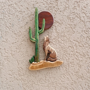 May include: Wooden wall art featuring a coyote howling at the moon, with a green cactus in the background. The moon is a brown circle with a wood grain texture.
