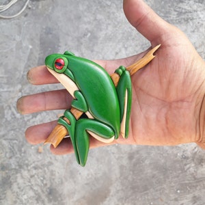 Puede incluir: Una rana de madera tallada a mano, pintada de verde con ojos rojos, posada en una rama. La rana se agarra a la rama con sus patas delanteras.