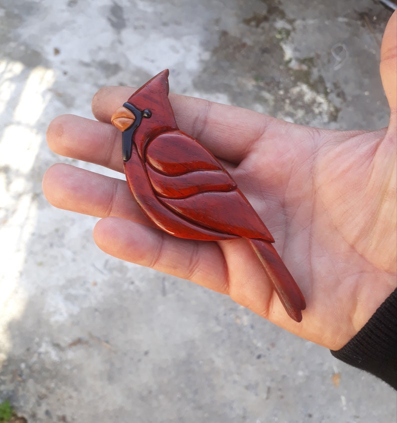 May include: A hand-carved wooden cardinal bird figurine with a red body, black eyes, and a brown beak. The bird is intricately detailed with wood grain visible.