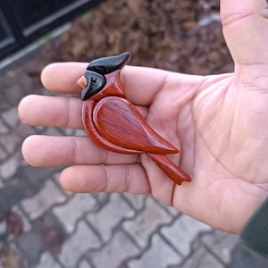 May include: A wooden cardinal bird figurine, hand-carved and painted. The bird features a vibrant red body, a black head, and a small, tan beak. The figurine is held in a hand, showcasing its size and detail.