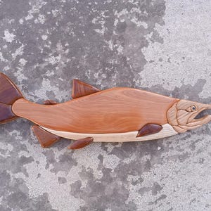 Puede incluir: Una escultura de madera de un salmón, tallada en tonos marrones y blancos. El pez está representado en una pose de salto, con la cola levantada y la boca abierta.