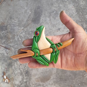 Puede incluir: Una escultura de rana de madera verde y blanca posada en una rama. La rana tiene ojos rojos y está tallada en un estilo realista.