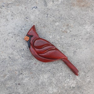May include: A hand-carved wooden cardinal bird figurine with a red body, black eyes, and a brown beak. The bird is perched on a gray surface.