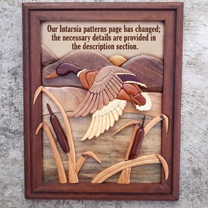 May include: A framed intarsia wood wall art piece featuring a mallard duck in flight over a marsh with cattails and hills. The text reads: Our Intarsia patterns page has changed; the necessary details are provided in the description section.