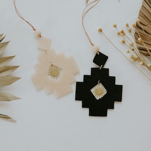 May include: Two decorative hanging ornaments. One is a light beige colour, and the other is black. Both have a geometric design with a sunburst pattern in the centre. They hang from a thin, light brown cord.