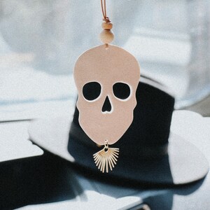 May include: A pink skull-shaped car air freshener with a gold sunburst charm hanging from a brown leather cord with wooden beads.