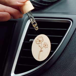 May include: A wooden car air freshener with a floral design. A hand holds a glass dropper bottle with a clear liquid dripping onto the air freshener.