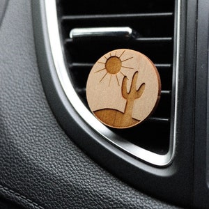 May include: A wooden car air freshener with a design of a cactus and a sun. The air freshener is attached to a car's air vent.