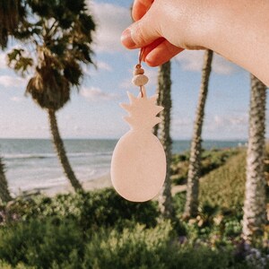 May include: A light pink pineapple-shaped car air freshener with a brown string hanging from the top. The air freshener is being held in a hand against a background of palm trees and a beach.