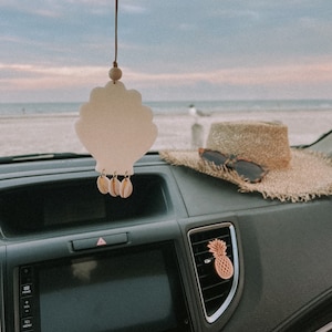 May include: A car interior with a seashell-shaped car air freshener hanging from the rearview mirror. The air freshener has small seashells hanging from it. There is a straw hat and sunglasses on the dashboard, and a pineapple-shaped air freshener in the air vent.
