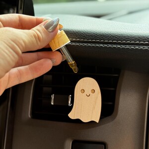 May include: A wooden ghost-shaped car air freshener with a smiling face is placed on a car vent. A hand holds a dropper with a yellow liquid, possibly essential oil, above the freshener. The car interior is visible in the background.