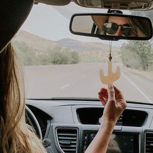 May include: Two people in a car, one wearing a brown hat and the other wearing a black hat. The person in the brown hat is holding a small, light brown cactus-shaped car air freshener.