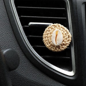 May include: A woven car air freshener with a seashell in the centre. The air freshener is beige and brown and is hanging from the air vent in a car.