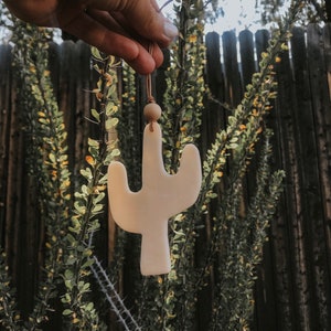 May include: A white ceramic cactus ornament with a brown leather cord and a wooden bead. The ornament is hanging from a hand.