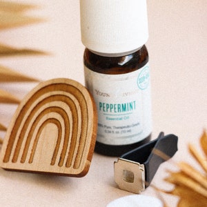 May include: A wooden rainbow-shaped car air freshener with a small bottle of peppermint essential oil, a black car vent clip, and a white cotton pad.