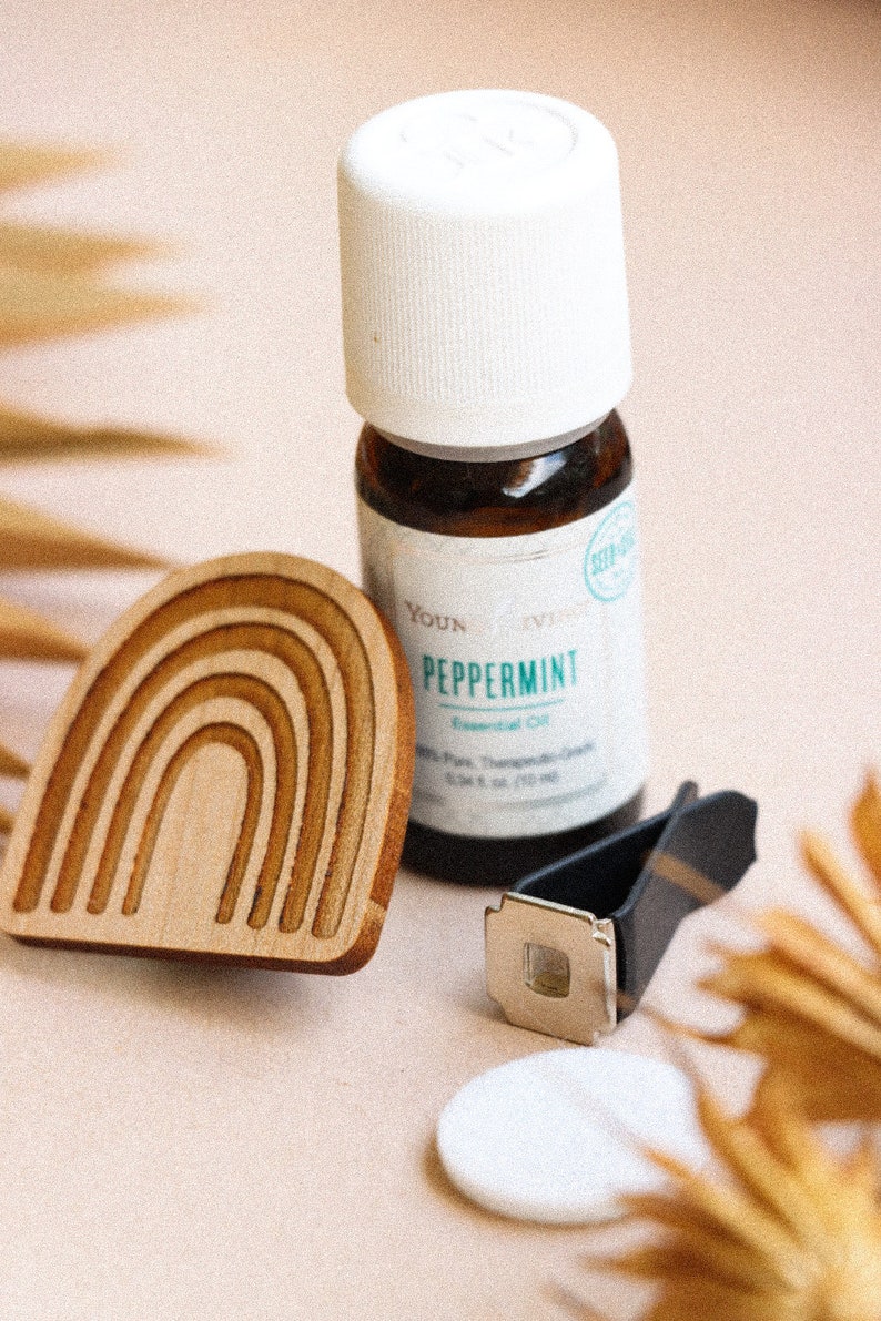 May include: A wooden rainbow-shaped car air freshener with a small bottle of peppermint essential oil, a black car vent clip, and a white cotton pad.