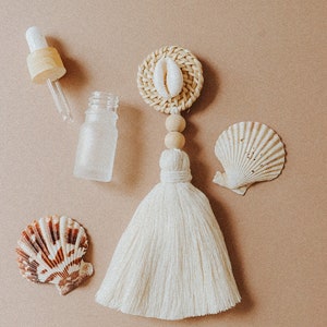 May include: A white tassel with a wooden bead and a woven circle with a white seashell. There are two seashells on a brown background, one brown and one white.