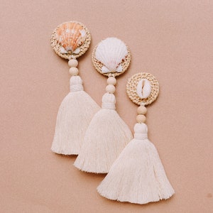 May include: Three white tassel ornaments with seashells and woven details. Each ornament has a different seashell and is attached to a wooden bead.