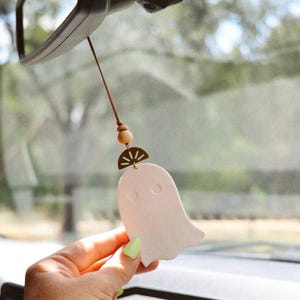 May include: A light pink ghost-shaped car charm with a brown cord, wooden bead, and gold-colored semi-circle accent. The charm is hanging from a car's rearview mirror, suggesting a Halloween or spooky theme.