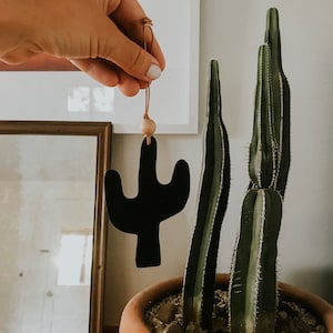 Peut inclure: Une décoration en forme de cactus noire suspendue à un cordon en cuir marron avec une perle en bois. La décoration est suspendue devant un cactus en pot.