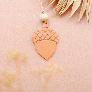 May include: A terracotta acorn ornament with a textured top and a smooth bottom. The ornament is hanging from a string with a wooden bead.