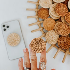 May include: A woven rattan phone grip and a matching phone case grip. The circular grips are light brown and tan. A collection of similar rattan coasters are in the background. The phone case is light gray.