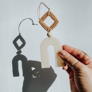 May include: A white ceramic arch-shaped pendant with a woven rattan square hanging from it. The pendant is held by a hand.