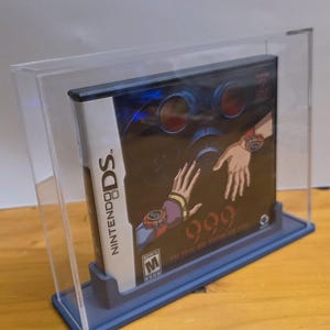 May include: A clear acrylic display case with a blue base, showcasing a Nintendo DS game cartridge. The game is titled "999: Nine Hours, Nine Persons, Nine Doors" and features a dark background with red and blue accents. The game is rated M for Mature.
