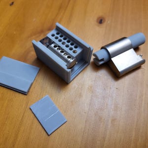 May include: A close-up of a gray plastic cabinet hinge with a silver metal latch. The hinge is disassembled and the parts are laid out on a wooden surface.