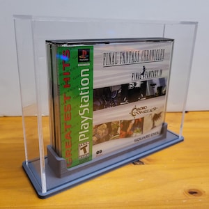 Op de afbeelding: Een transparante acryl displaykast met een grijze basis, die een PlayStation-spelcartridge toont. De game is Final Fantasy Chronicles, met Final Fantasy III en Chrono Trigger. De gamecase is gelabeld met "Greatest Hits" en "PlayStation".